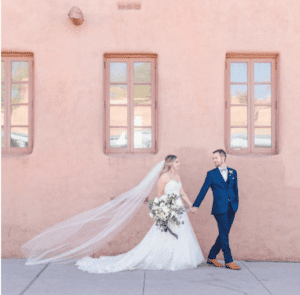 Woman in wedding dress holding flowers and man in blue suit walk in front of pink wall